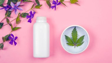 Pembe arka planda kozmetik ürünleri olan beyaz şişeler marihuana yaprağı ve mor kır çiçekleri manzaralı. güzellik, cilt bakımı