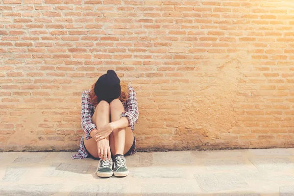 Young woman sad sitting against brick wall alone.