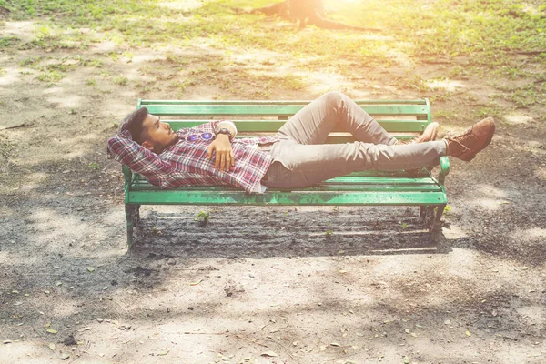 Young hipster man lying down on green park bench, looking away.