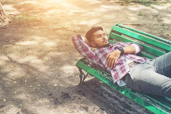 Young hipster man lying down on green park bench, looking away.