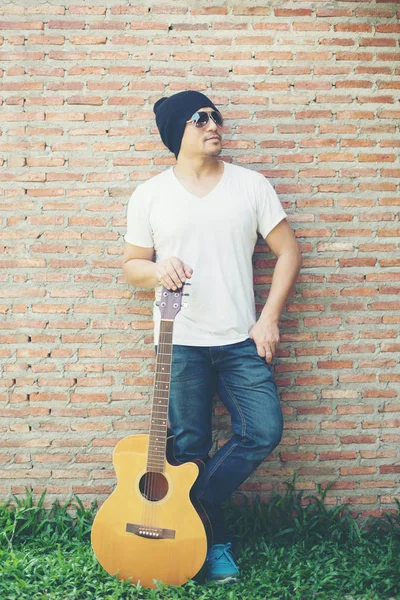 Handsome guy standing holding guitar against the brick wall posi — Stock Photo, Image
