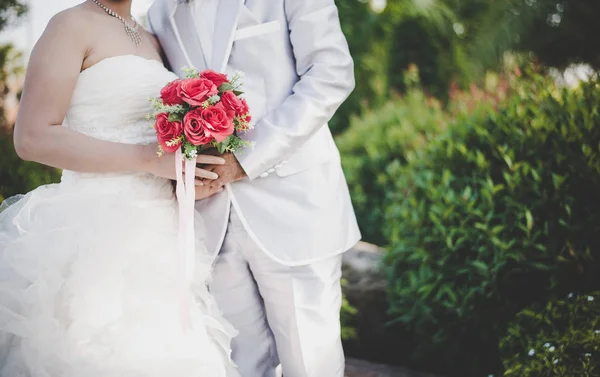 Bruden håller en röd ros brudbukett i händerna, brudgummen kramar — Stockfoto