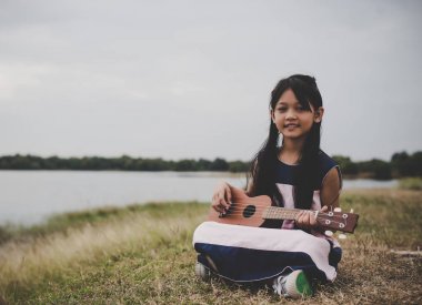 Sevimli küçük Asya Kız parkta çim ve oyun ukulele üzerinde oturan.