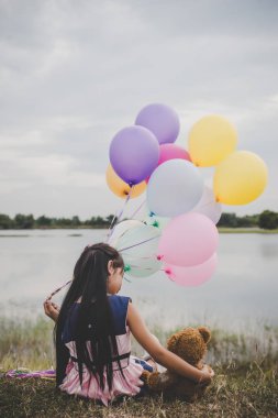 Oyuncak ayı dışında uzun yeşil çimenlerin üzerinde oturan küçük şirin kız. Renkli balonlar elinde tutan kız. 