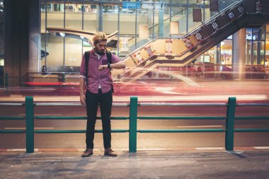 Hipster sakallı adam ayakta backpacker seyir smartwatch geceleri seyahat ederken araba hareket arka plan ile.