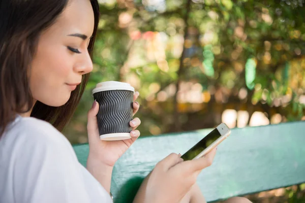 Portrait of a happy casual woman sitting on a bench with coffee and looking on the phone in the park. Women lifestyle concept.