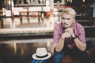 Tren istasyonunda sırt çantası ile tahta bankta oturan genç hipster adam. oturan adam tren platformu için bekliyor.