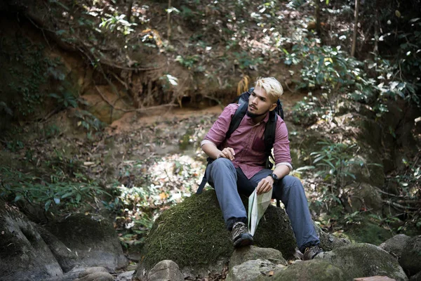 Young tired hiker with backpack sitting on the rock in forest while resting after active walk. Travel concept