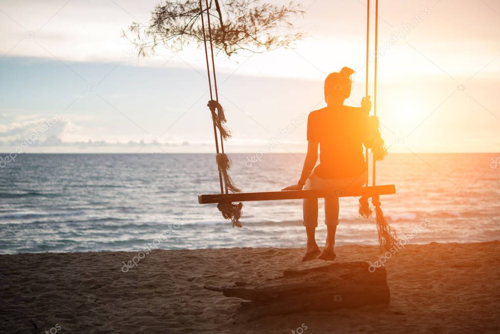 Young woman watching sunset alone sitting on swings on the beach at sunset