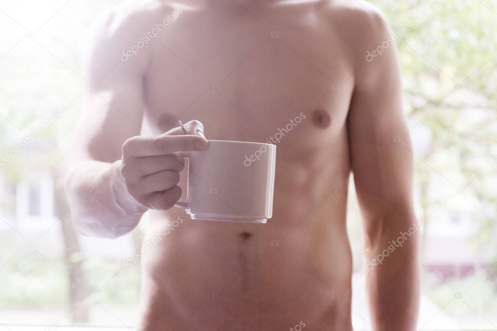 A young man holding a mug in his hands. Morning, summer, autumn, bare-chested. Tea, coffee.