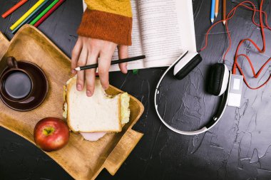 Biri okur, metni çevirir. Sandviç, kulaklık, kalem, defter. Natürmort, siyah arka plan.