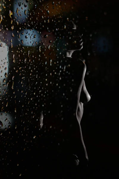 Silueta femenina atractiva, contorno sobre fondo oscuro bajo gotas de lluvia.Vista a través de la ventana, vidrio — Foto de Stock