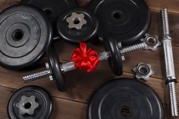Hediye: Koyu ahşap bir masada kırmızı kurdeleyle bağlanmış halterler ve spor malzemeleri.