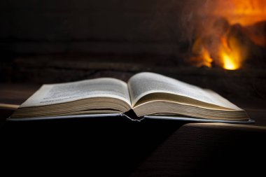 Açık bir kitap yanan bir şöminenin yanındaki bir ahşabın üzerinde yatıyor.