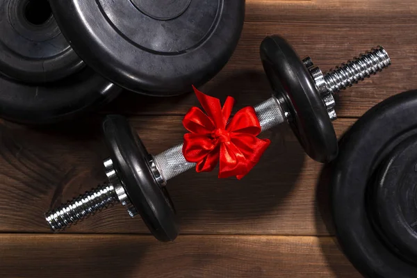 Hediye: Koyu ahşap bir masada kırmızı kurdeleyle bağlanmış halterler ve spor malzemeleri.
