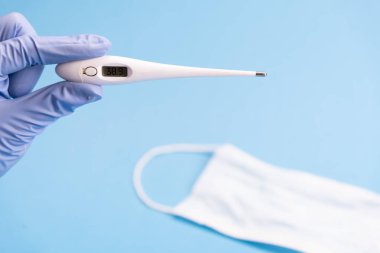 Doktorun elinde tıbbi maskeli mavi arka planda dijital bir termometre var. Yakın plan. Tıbbi tesis