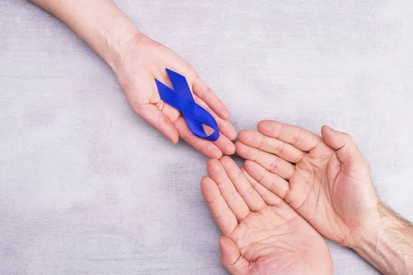 Una mujer sostiene una cinta de conciencia azul, símbolo en las manos masculinas, cáncer de colon, cáncer colorrectal sobre un fondo gris con espacio para texto, posición plana, vista superior — Foto de Stock