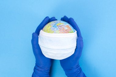 Küredeki tek kullanımlık tıbbi maske mavi arka plandaki bir doktorun elinde, Dünya 'nın modeli. Salgın konsepti. KOVID-19, koronavirüs. Hastalığın küresel salgını. COVID Tehlikeli Virüs
