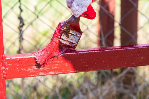 Hands in construction gloves paint a metal fence with red paint.