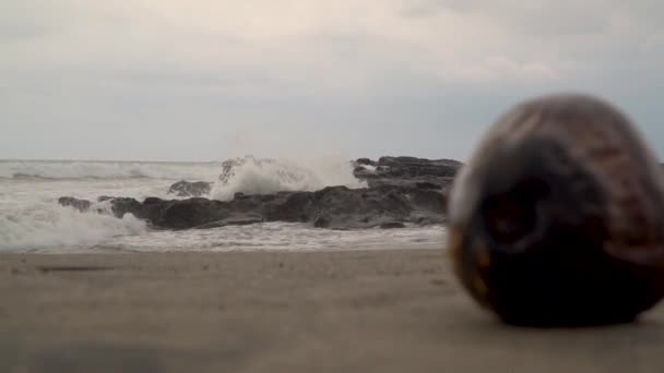 Vista del océano y olas rompiendo piedras y rocas sobre arena yace un coco — Vídeo de stock