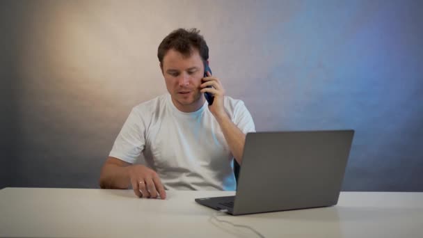 Un hombre se sienta en una mesa con el ordenador portátil y hablando por teléfono — Vídeos de Stock
