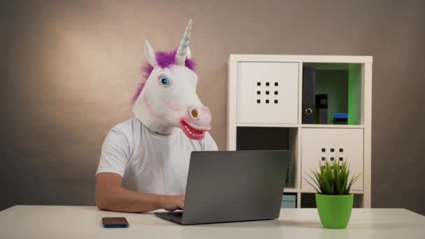 Человек в маске единорога сидит за столом и работает над ноутбуком — стоковое видео