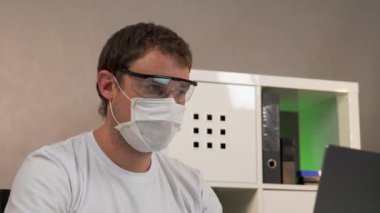 Adam koruyucu gözlük ve maskeyle dizüstü bilgisayarda çalışıyor.