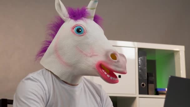 El hombre en la máscara de un unicornio sentado en una mesa de trabajo en el portátil — Vídeo de stock