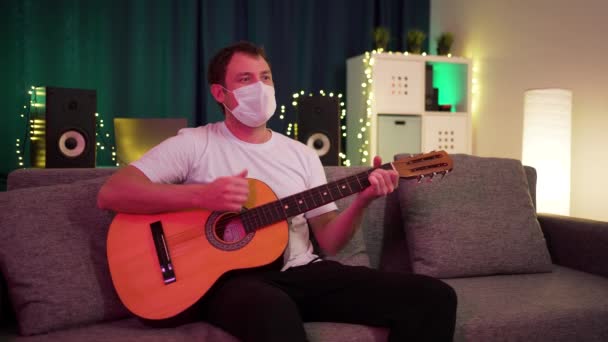 Мужчина играет дома на гитаре в защитной маске — стоковое видео