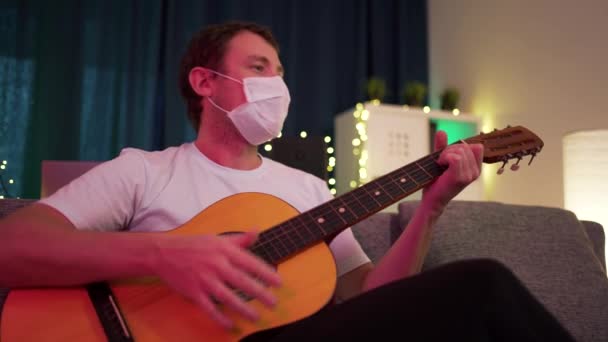 Een man speelt thuis gitaar met een beschermend masker — Stockvideo