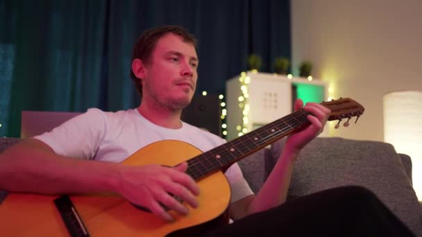 Un hombre toca la guitarra sentado en el sofá — Vídeos de Stock