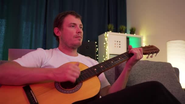 En man spelar gitarr sittandes på soffan — Stockvideo