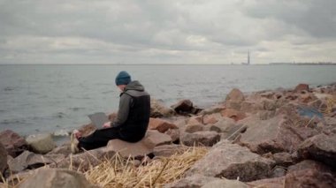 Maskeli adam. Körfezin kıyısında dizüstü bilgisayarlı bir kayanın üzerinde oturuyor.