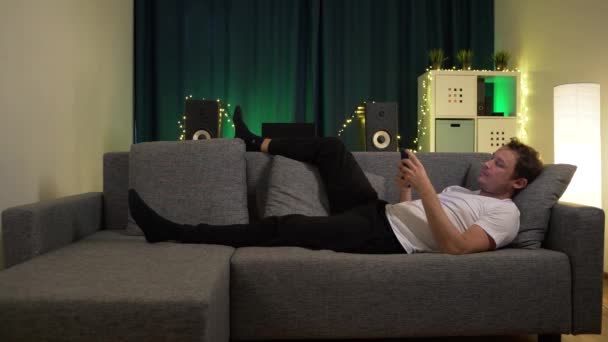 En man ligger på soffan, med telefonen — Stockvideo