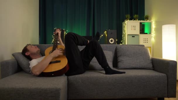 Een man speelt gitaar terwijl hij op de bank ligt — Stockvideo