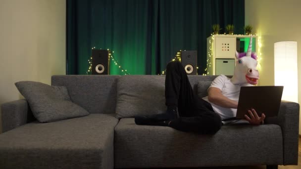 Hombre en la máscara unicornio sentado con el ordenador portátil en el sofá y está escribiendo un mensaje — Vídeo de stock