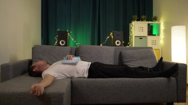 Um homem deitado no sofá com um livro no peito, e dormindo — Vídeo de Stock