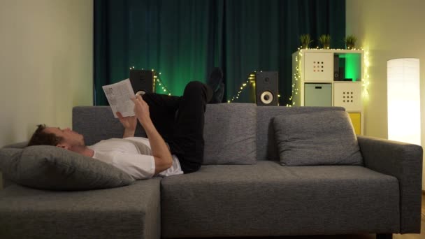 Ein Mann liegt auf dem Sofa und blättert in einem Buch — Stockvideo