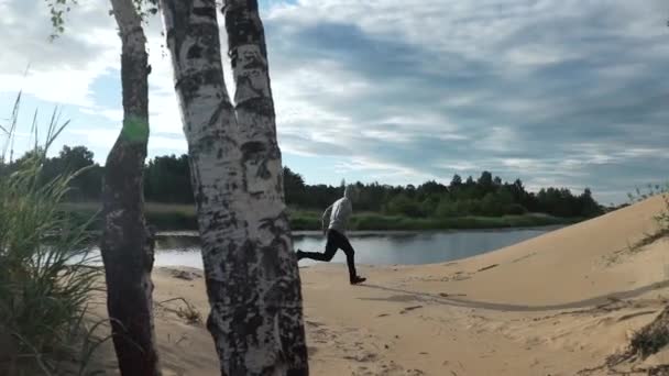 Muž běží pískem podél jezera šplhá na kopec — Stock video