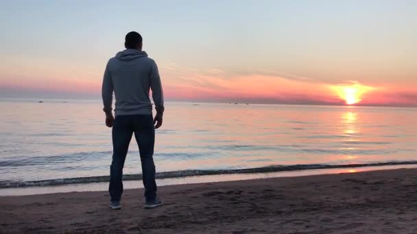 Ένας άντρας στέκεται στην παραλία το ηλιοβασίλεμα απολαμβάνοντας τη θέα — Αρχείο Βίντεο