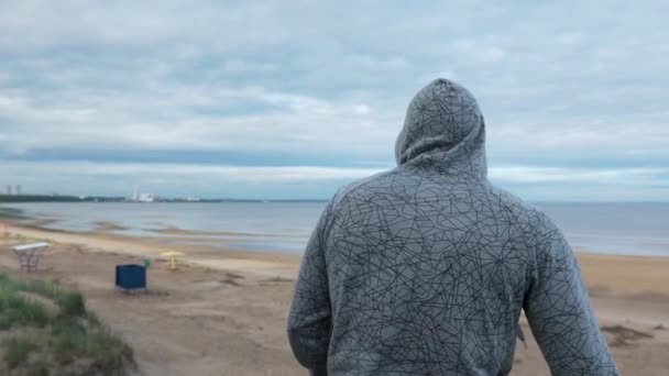 Un hombre va a la playa y se quita la capucha — Vídeo de stock