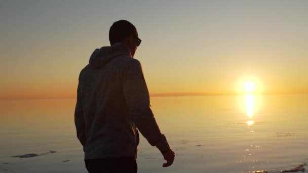 Man in zonnebril is op het strand langs de zee bij zonsondergang — Stockvideo