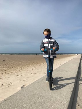 Covid-19 döneminde yüzünde siyah bir maskeyle Baltık sahilinde scooter süren genç bir çocuk..