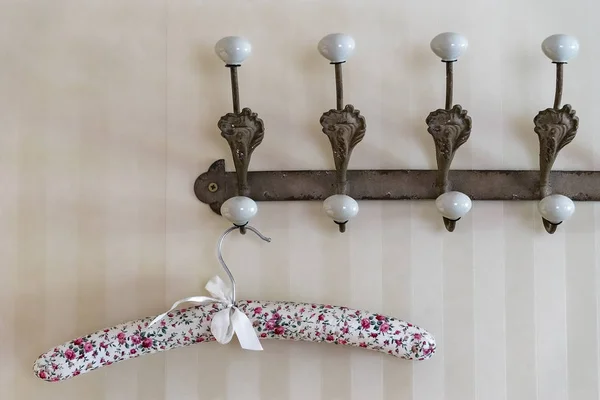 Stylish floral cloth covered clothes hanger hanging on a brown vintage wall hanger with white hooks. The hanger is decorated with white ribbon. Light yellowish striped background
