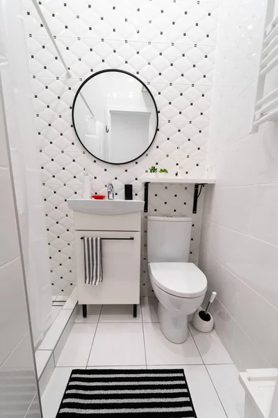 Part of a small new modern bathroom. The main colors - white and black. Tiled walls and floors. Cabinet with sink, shelf and mirror on the wall, toilet, toilet tank, a rug on the floor