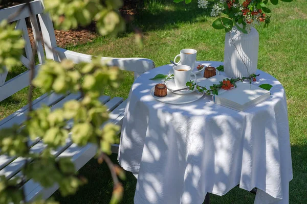 White tablecloth covered table with coffee, cakes, book and vase in the garden by a white bench behind a tree branch with fresh green leaves. The concept of relaxation. Selective focus on the table