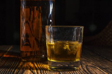 Bir bardak buzlu viski, ahşap bir masada bir şişe. Işıklı eski bir masa üstü ve bir bardak sert içki..