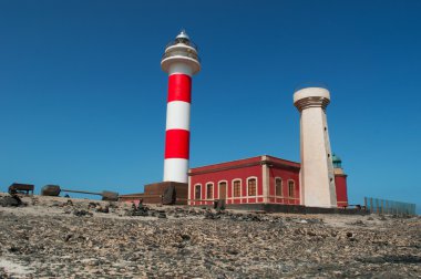 Fuerteventura, Canary Islands, Spain: black rocks and view of Faro de Toston (Toston lighthouse or El Cotillo lighthouse), an active lighthouse at Punta de la Ballena (Whale Point) whose original structure was opened in 1897 clipart