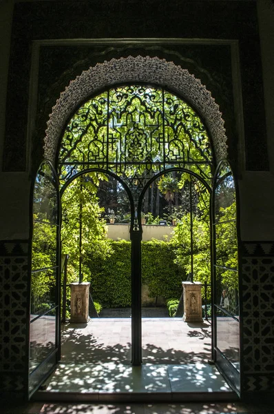 España: los jardines del Alcázar de Sevilla, el famoso palacio real de la ciudad, destacado ejemplo de arquitectura mudéjar, visto a través de una puerta de arco de hierro forjado — Foto de Stock