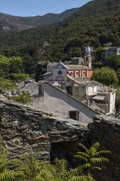 Córcega: la iglesia pintada de rosa de Santa Julie (siglo XVI) en el horizonte de Nonza, pueblo en la costa occidental de Cap Corse con casas antiguas encaramadas en los acantilados sobre 100 metros del mar — Foto de Stock